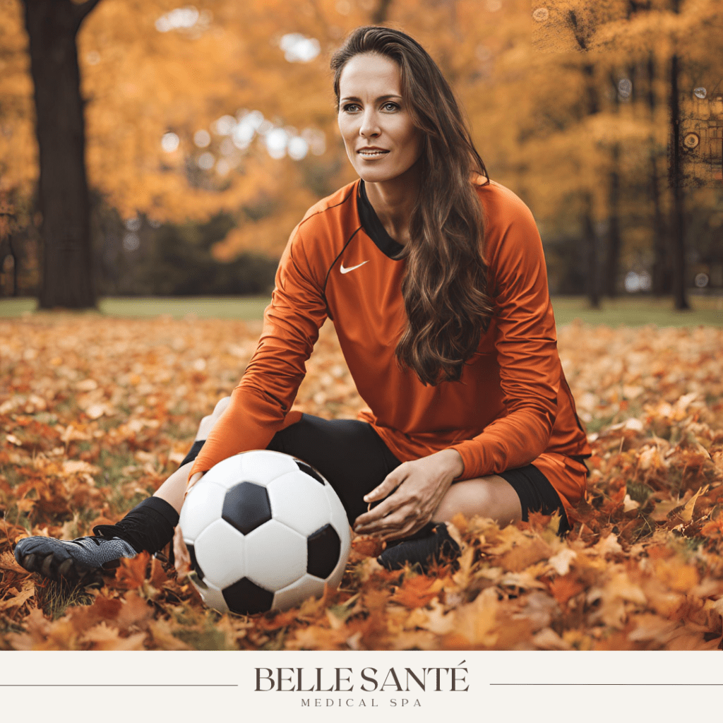 Woman in soccer uniform holding a soccer ball on the ground with a background of Fall leaves.